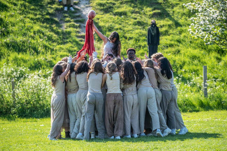A group of young people huddled in a circle, holding one girl up in the middle. She is holding a red rag aloft. Everyone is wearing beige clothing. They appear to be in a park.