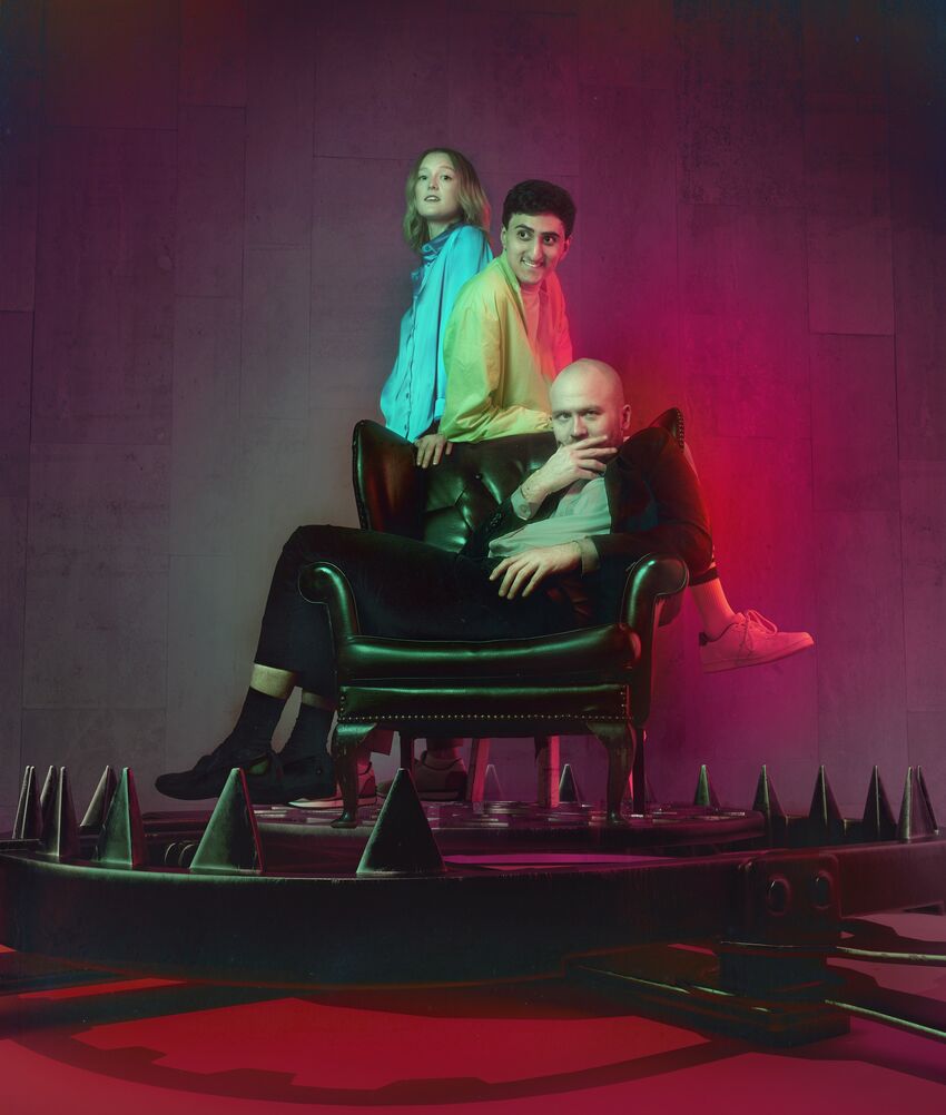 A man reclines in a leather armchair with his hand thoughtfully over his mouth. Another man leans on the back of the chair smiling. A woman stands behind him with a hand on the chair looking defiantly at camera. Captioned and Audio Described icons