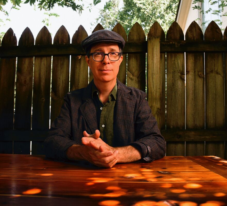 Ben Folds sitting on a wooden table with a fence behind him. He has his hands to together