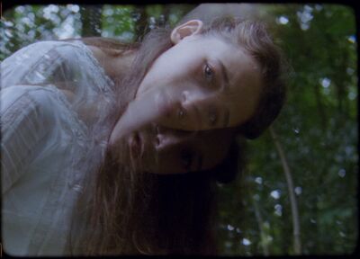 A young woman in a white dress looks directly down the camera. Using a double exposure effect the image is slightly blurred and gives the feeling of motion