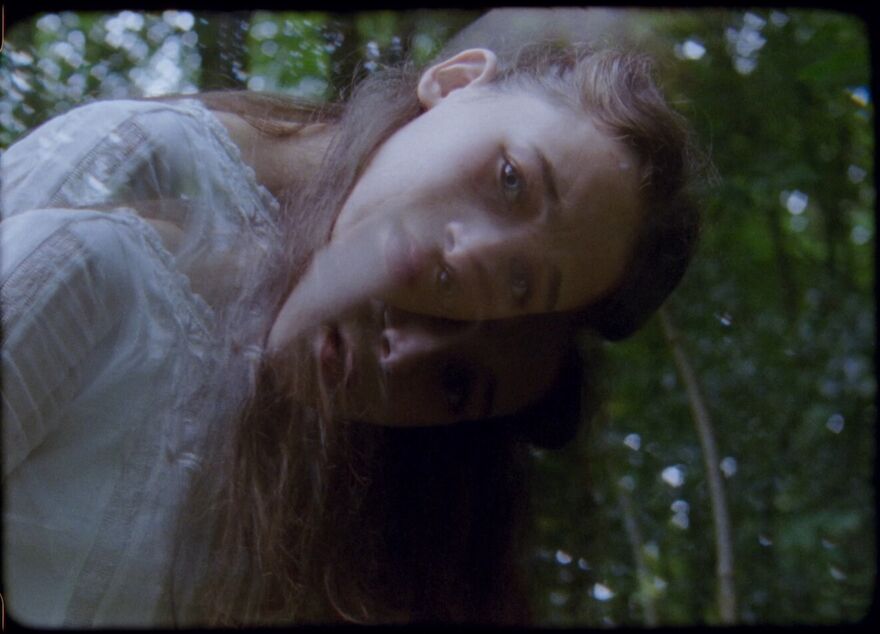 A young woman in a white dress looks directly down the camera. Using a double exposure effect the image is slightly blurred and gives the feeling of motion