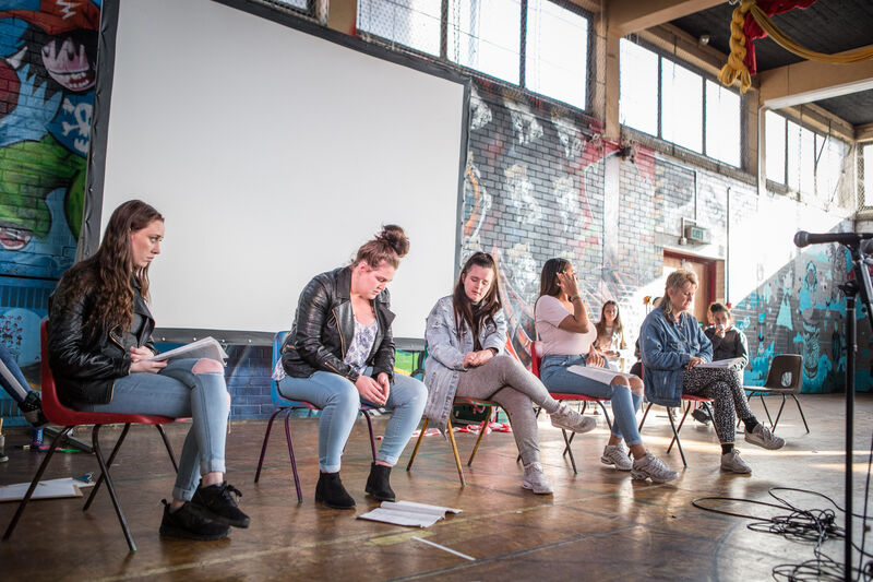 Five young people are siting on chairs in a row against a graffitied wall and projector screen