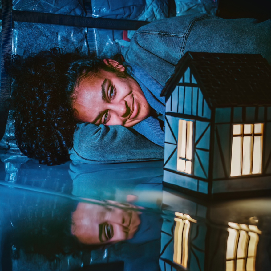 A girl lays on the floor, her head resting on her bent arm. She looks at a model of a house that has the lights on, glowing inside