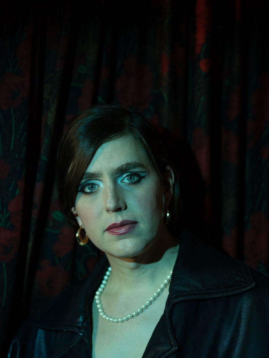 Photograph of Ezra Furman from the shoulders up wearing a pearl necklace, gold hoop earrings and a black leather jacket against a black urtain