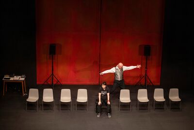 Tim Crouch moving across a stage with his arms stretched apart, a row of chairs in front of him with Jessie Buckley sitting forwards on one
