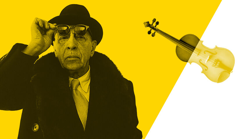 A man wearing a black coat and hat holds up one hand to lift some sunglasses from his face. To his left is a violin