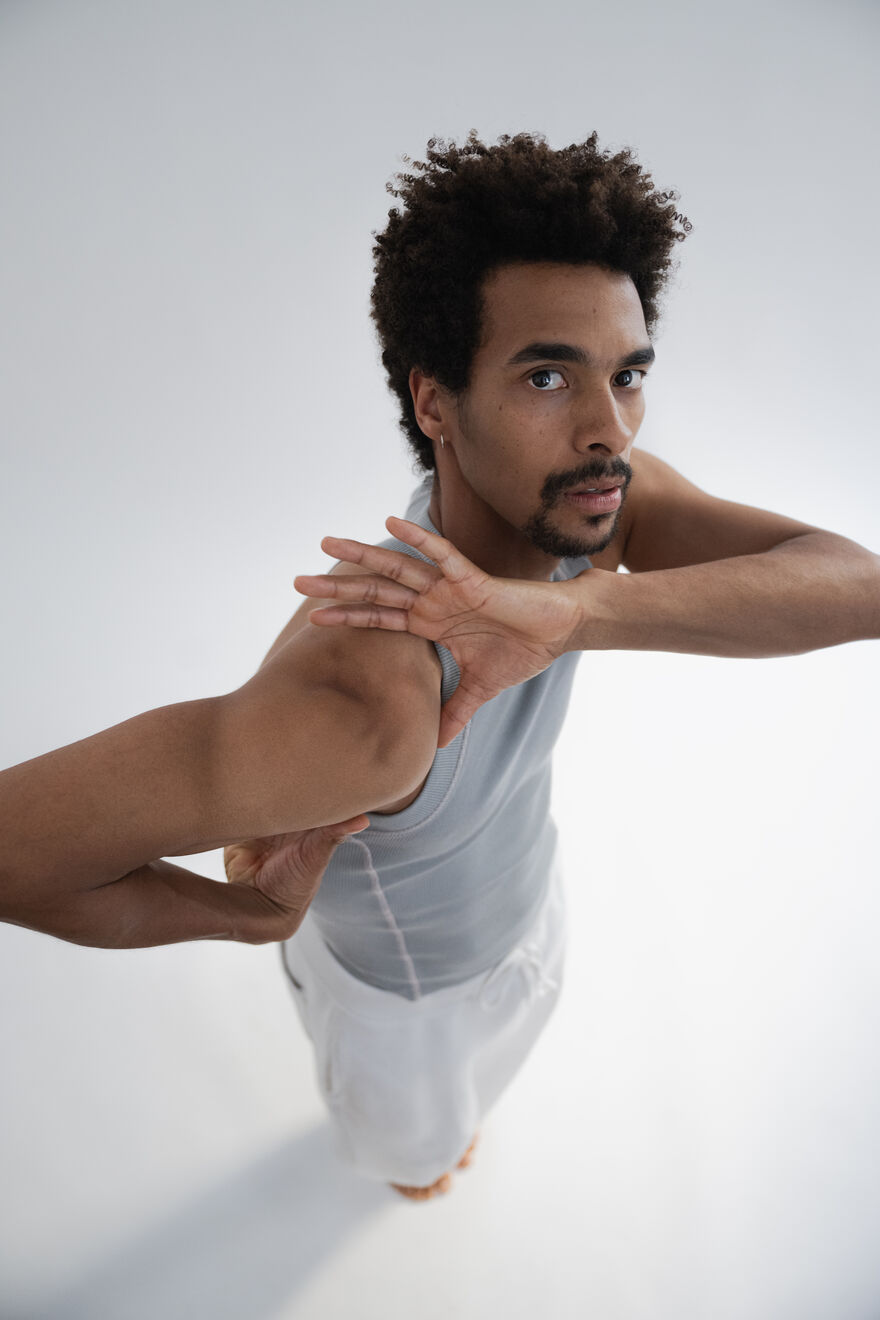 A man in a grey tank top and white trousers, staring at the camera with one arm behind his back and one on his shoulder