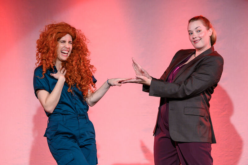 Two performers are on stage, one has a red curly wig and the other a business jacket holding open a fake box mirroring a scene from Pretty Woman