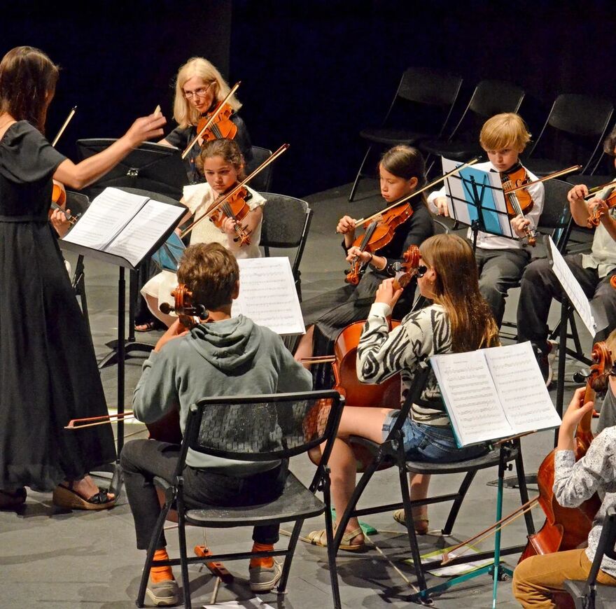 Image of a group of children playing violins on stage being conducted