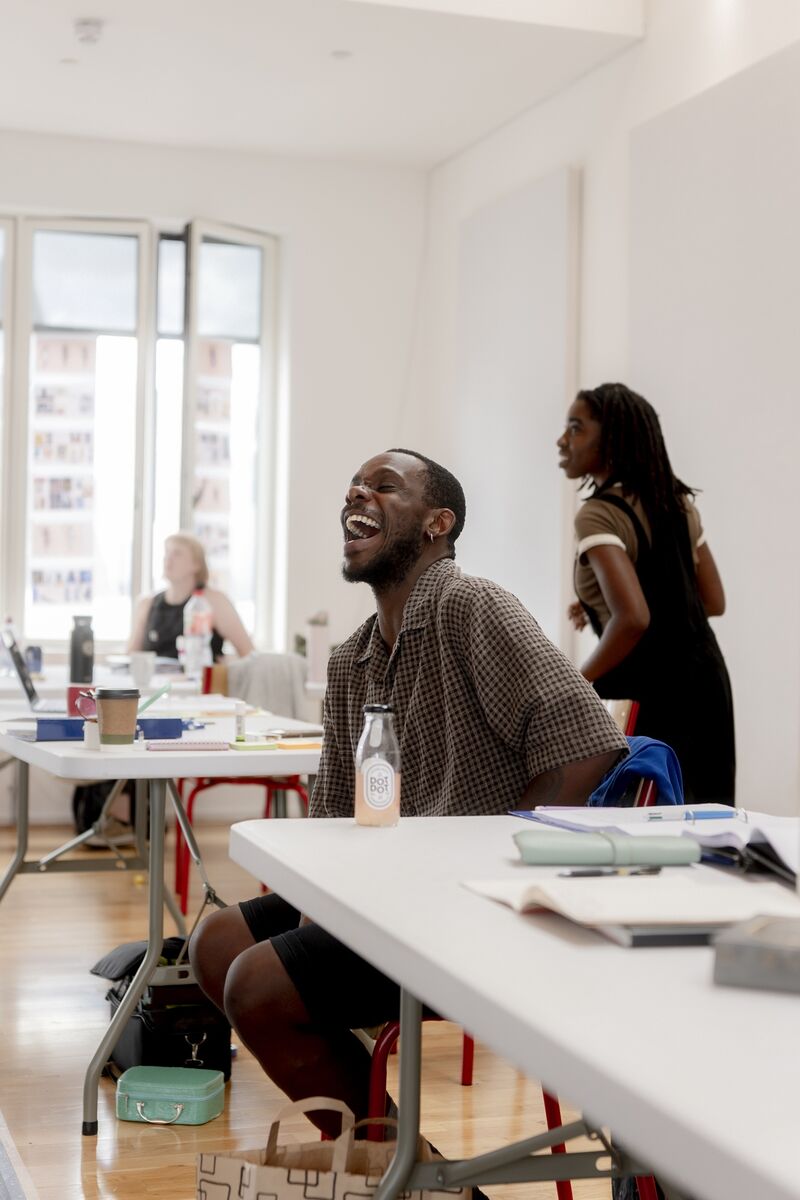 A black man sat in a chair is laughing, his mouth open wide with joy!