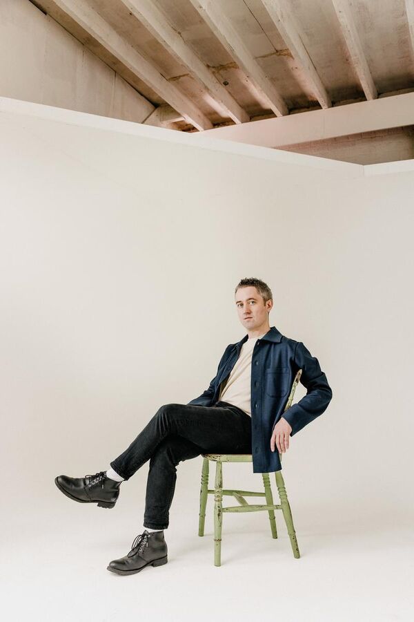 Conor O'Brien sat on a wooden chair wearing casual clothing in a white photography studio