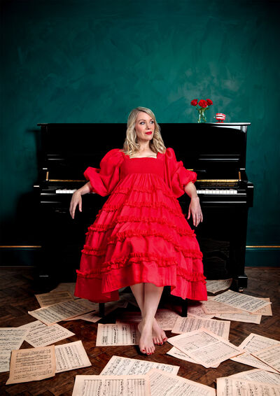 Rachel Parris wearing a red dress leaning on a piano with music sheets strewn on the floor around her feet