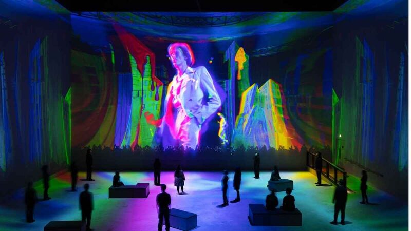 An immersive projection featuring a brightly lit figure against colorful, abstract cityscape imagery, with visitors silhouetted in the foreground.