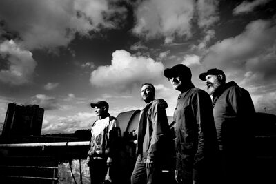 Black and white image of four men walking. They are walking in front of a cloudy sky.