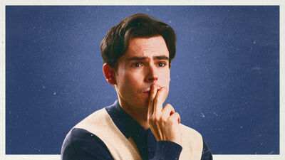 Rhys James in front of a blue background looking away from the camera with two fingers to his mouth