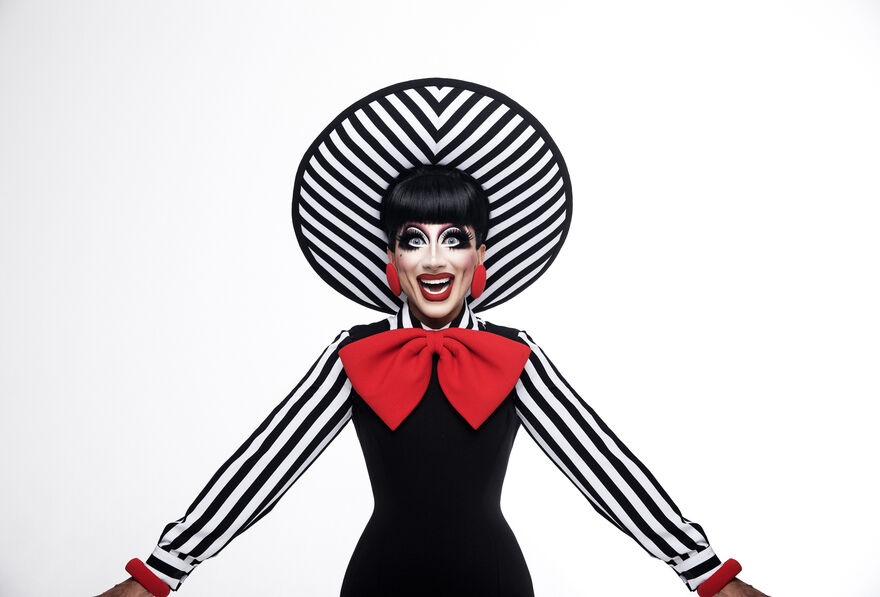 Bianca De Rio wearing a black and white stripey dress with a red bow. They have a happy facial expression and a stripey hat too.