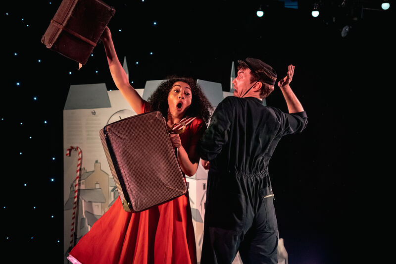 A girl in a red sailor dress holds a suitcase in each hand looking as though she's about to stumble while a man backs away