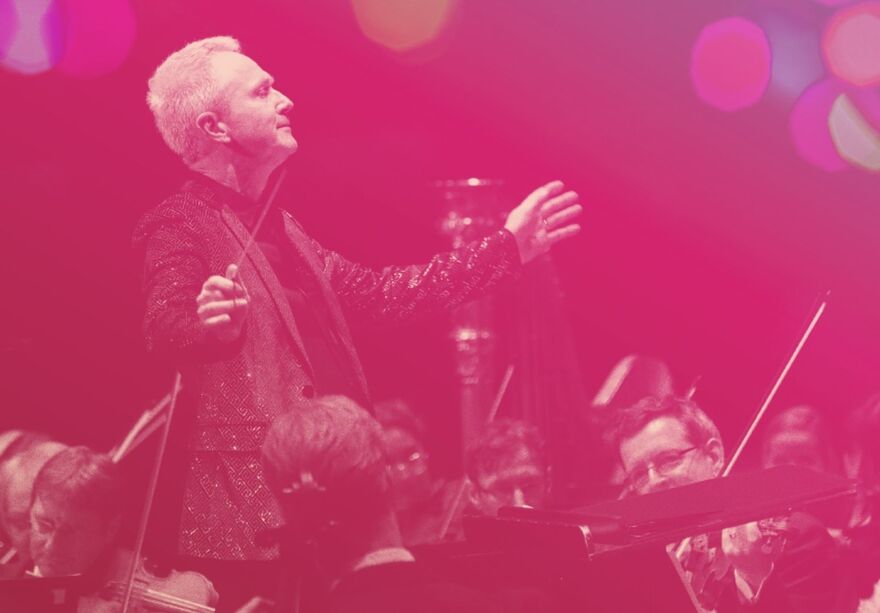 Conductor holding up his arms in front of an orchestra. The photo is treated with a pink haze and purple bubbles