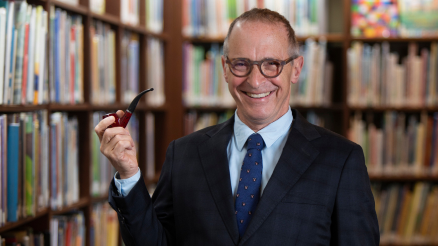David Sedaris surrounded by bookshelves wearing a suit, glasses and holding a smoking pipe