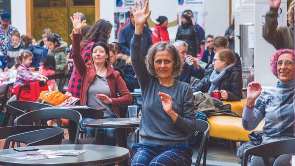 A group of people are sat in the Brighton Dome Foyer. They have their hands in the air following someone