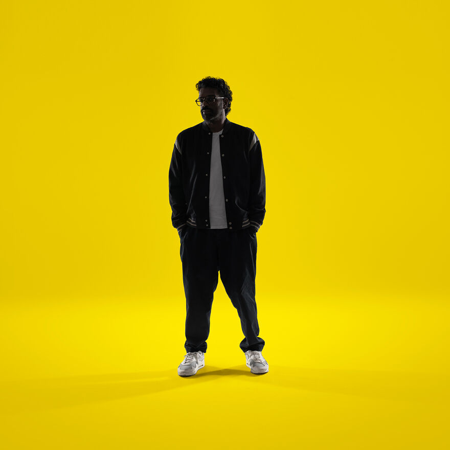 Romesh Ranganathan standing with his hands in his pockets over a yellow background