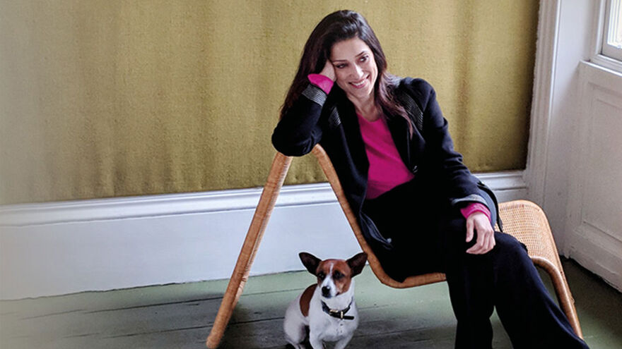Fatima Bhutto sitting sideways on a chair, wearing a suit and pink shirt, with a dog sitting to the left of her
