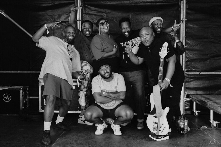 The band BCUC standing and crouching staring at the camera. Some members are holding guitars, crossing their arms or doing the peace sign. The image is in black and white