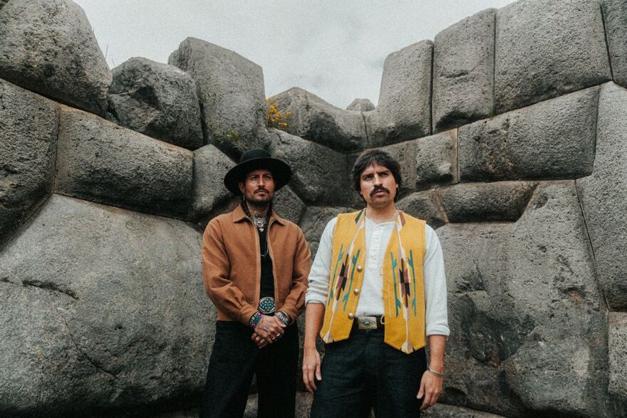 Two men pose in front of massive stone walls, wearing colorful patterned clothing.