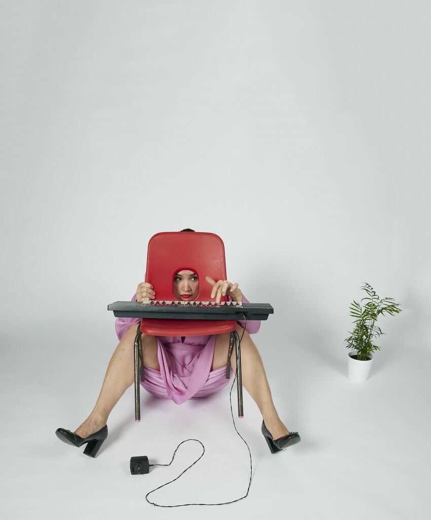 Rachel Blackman sitting on the floor behind a red plastic chair, with her legs on either side of the chair and her head through the hole in the chair's back. There's a small electric keyboard on the chair and a pot plant next to it
