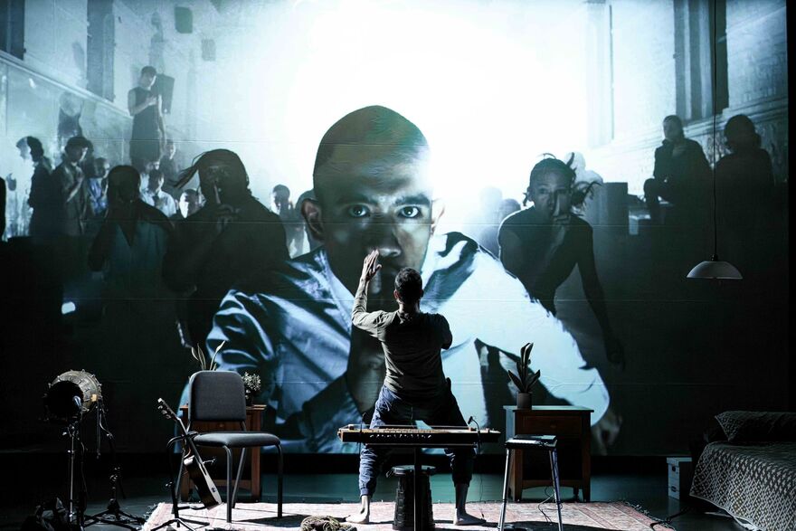 A man stands on stage facing away from the audience, he is stood in front of a screen showing people running towards him. On stage there are some chairs and instruments set up behind the man.