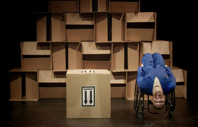 A man is upside down in his wheelchair. He is in front of a wall made from cardboard boxes and next to him is another cardboard box with two down arrows on it.