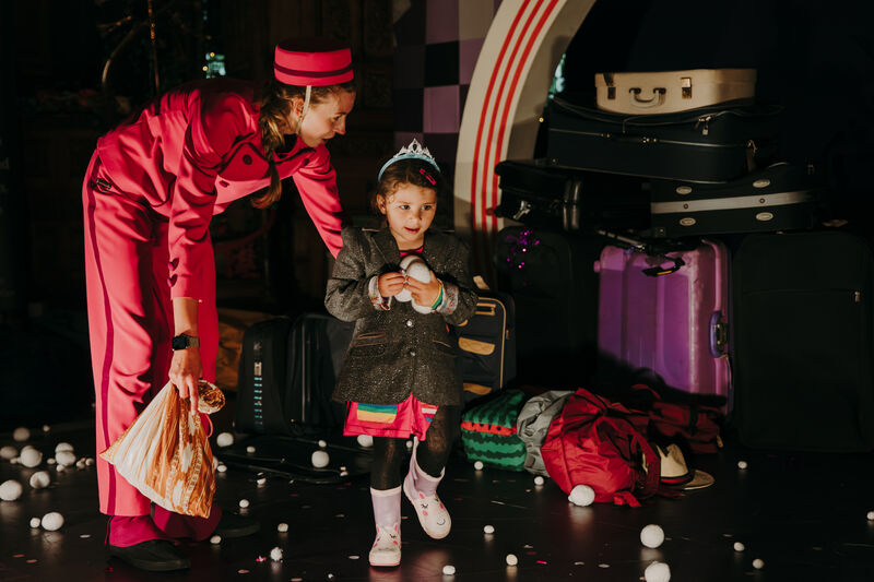A woman in a red jumpsuit and hat is leaning down towards a young child who is wearing a gray jacket and a tiara. The child is holding a white object and smiling. There are several suitcases on the stage