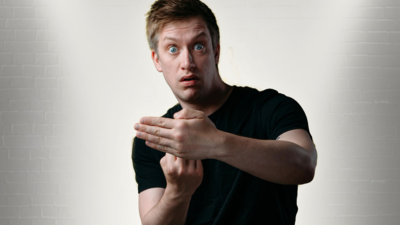 man in a black shirt in front of a brick white wall looks directly forwards with a surprised expression