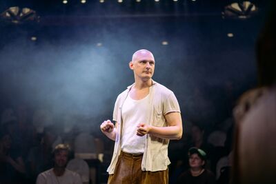 A man in a white top and cream shirt on a stage. There is haze and spotlights on him