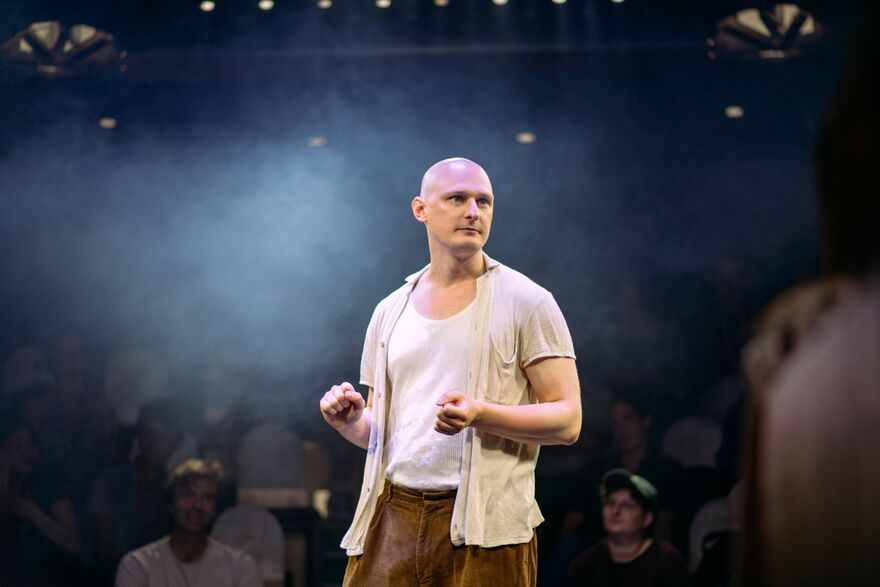 A man in a white top and cream shirt on a stage. There is haze and spotlights on him