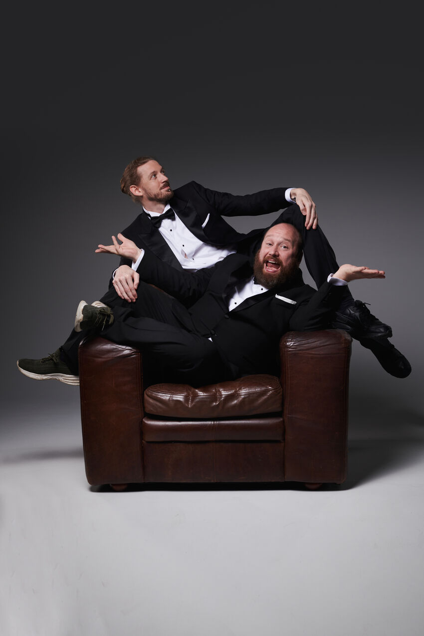 Two white men in black tuxedos sit on a brown leather chair, one has their legs off to the right hand side, the other to the left hand side.