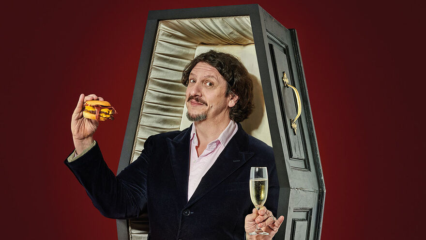 Jay Rayner wearing a suit standing in an open up-right coffin holding a burger in his right hand and a champagne flute in his left, with a deep red background
