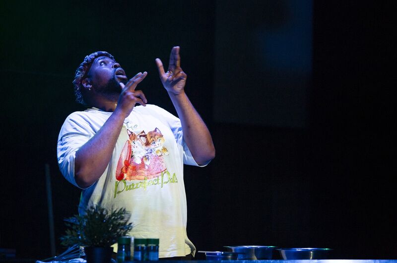 A black man in a white t-shirt doing finger guns up towards the ceiling, he is lit with a blue light.