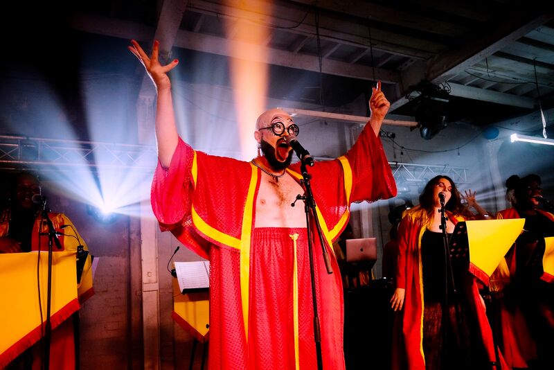 Fatt Butcher stands in the middle of the stage wearing a red and yellow robe, hands up to the sky with a choir singing in the background