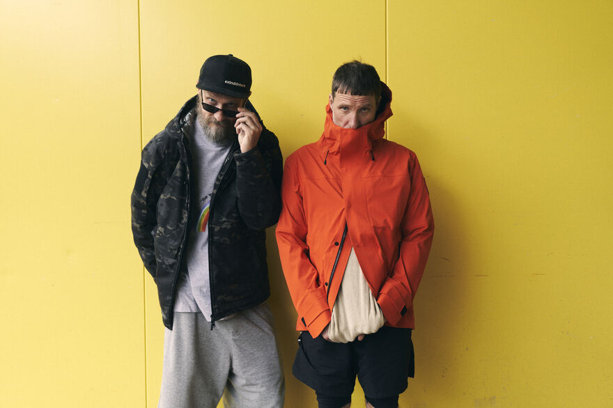 Two men wearing coats standing against a yellow wall. The one on the left is wearing a cap and sunglasses, which he is pulling down, and the one on the right is wearing an orange coat covering his month