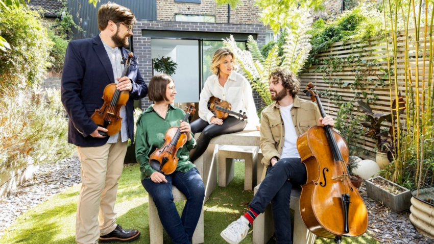 four people are in a garden looking at one another holding their instruments