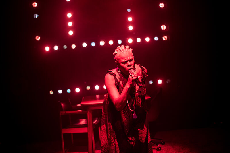 Sutara Galye on stage lit by red lights. She holds a microphone and is singing passionately