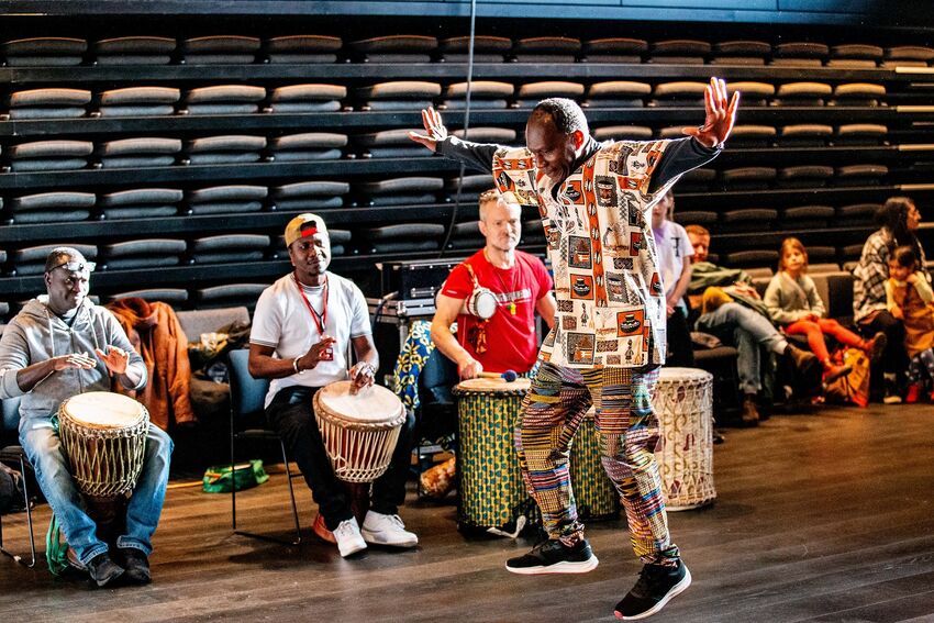 A row of Djembe drummers sit playing their drums whilst a man in colourful clothing dances in front of them