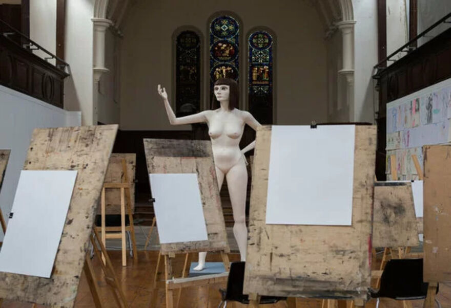 A mannequin of a tall woman with black hair stands in the middle of a church, around her are easels with blank white paper on them
