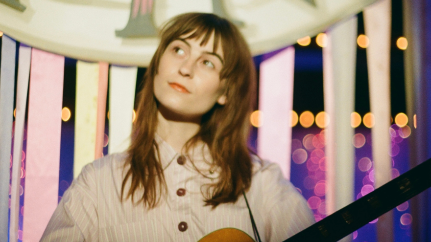 Faye Webster on stage holding an acoustic guitar looking thoughtfully into the distance. She is a white woman in her 20s with long brown hair wearing a casual buttoned-up shirt