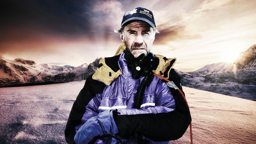 man in a coat and a hat situated in a mountainous and cold environment looks directly forwards 