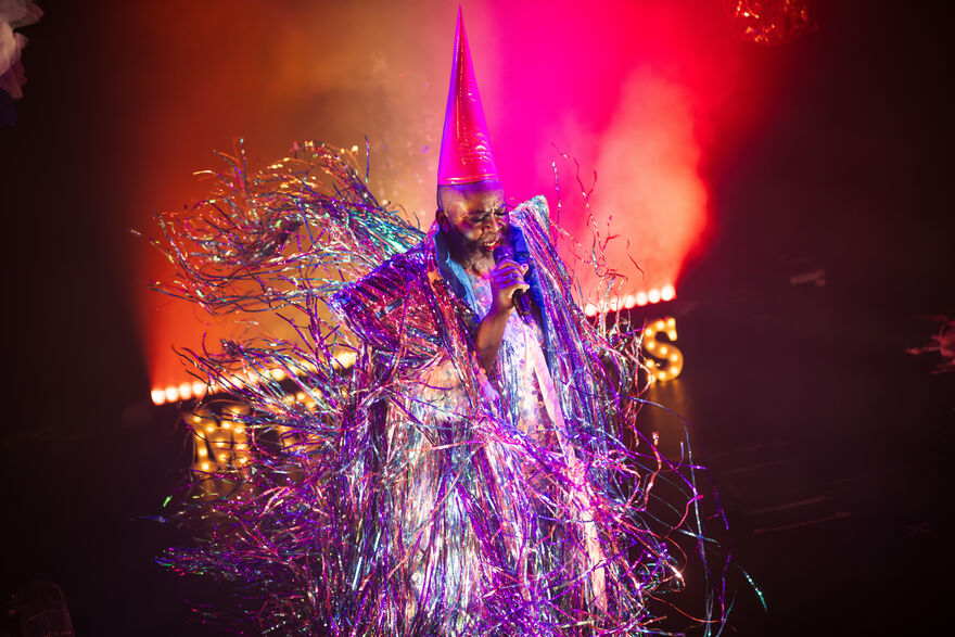 A person standing on a stage singing into a microphone. They are wearing a purple, pink tinsel outfit which is blowing behind them, they are also wearing a pink pointy hat.