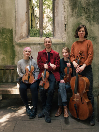 Group of string musicians sitting in an old building. They are said on a bench, holding violins, violas and a cello