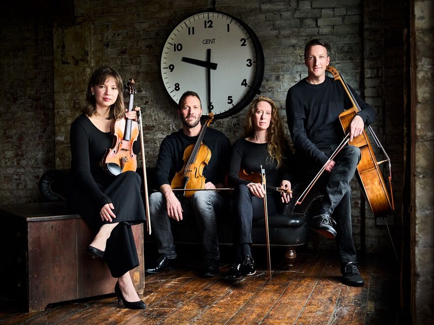 Four members of the Heath Quartet sitting on vintage furniture in an industrial-style room with bare brick wall and exposed wooden flooring. There is an oversized clock on the wall behind them showing the time at half past nine. They are lit by natural light from a window that's out of shot on our right hand side. From left to right, there is a lady with shoulder-length brown hair holding a violin and wearing a plain black long-sleeve top, black trousers and black heels. Next to her is a man with short dark hair and facial hair, he is also holding a violin and wearing all black long sleeve top, jeans and black shoes. Next to him is a woman with long wavy mousey-brown hair. She is wearing a plain black long-sleeve top and trousers with black shoes and also holding a violin. Next to her is a main with short dark hair, he is wearing a plain black long-sleeve top and black jeans and shoes. His cello is resting against his side.
