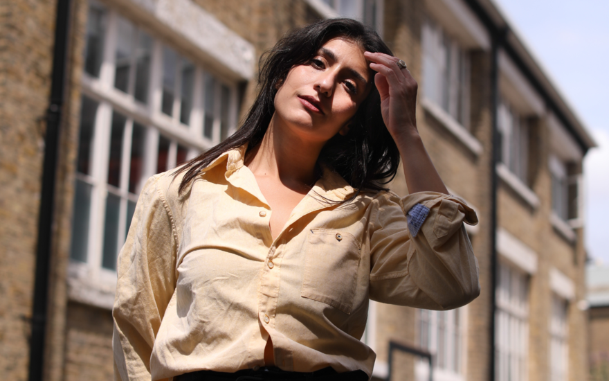 A woman standing in a sunny street wearing a cream shirt, with her hand on her hair